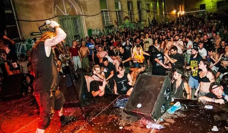 A high-energy night photograph of a live hardcore punk performance at the 25th Monteparadiso Festival in Pula, 2017. A vocalist with long hair leans from the stage into a dense crowd actively moshing in the courtyard of the Karlo Rojc social center